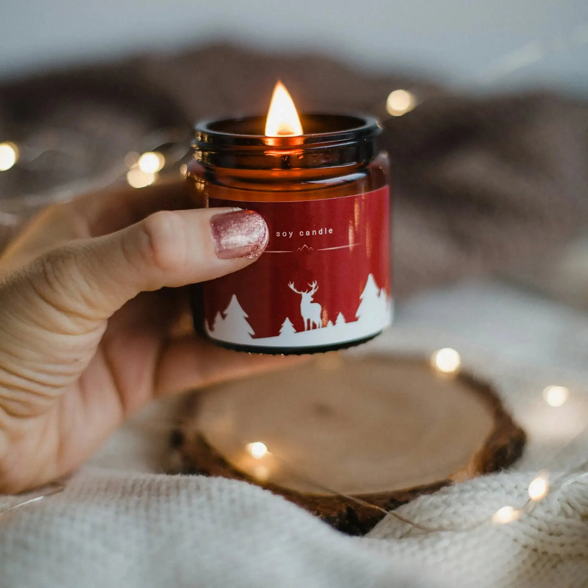 Hand holding a lit soy candle in an amber jar with a red Christmas label, white trees and a deer, against blurred fairy lights.