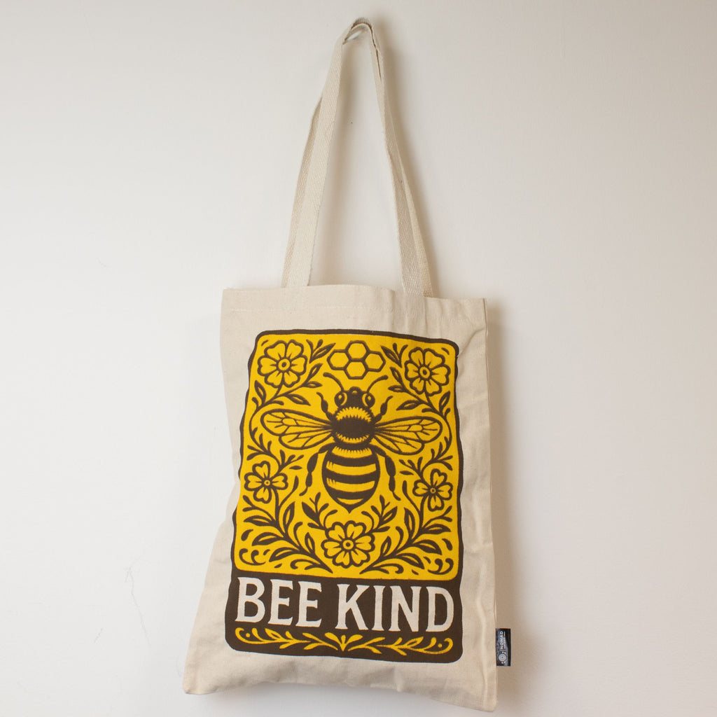 Beige tote bag with a bee and floral design, featuring the text 'BEE KIND', on a light background.