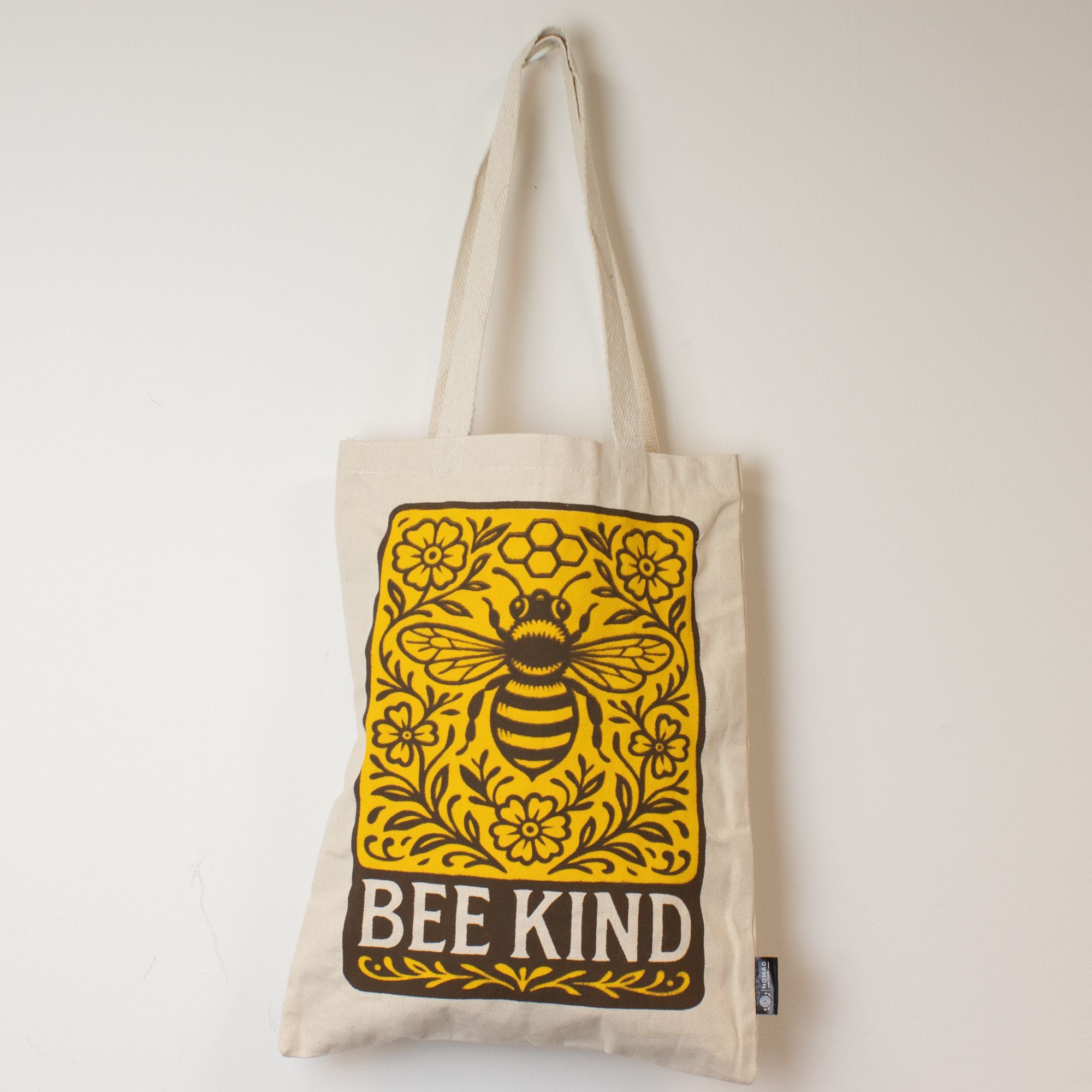 Beige tote bag with a bee and floral design, featuring the text 'BEE KIND', on a light background.