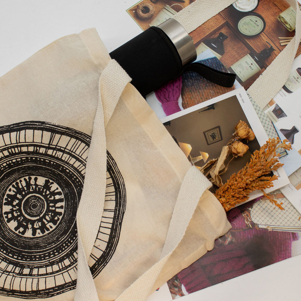Beige tote bag with black pattern on a surface with various items including a camera and dried plants.