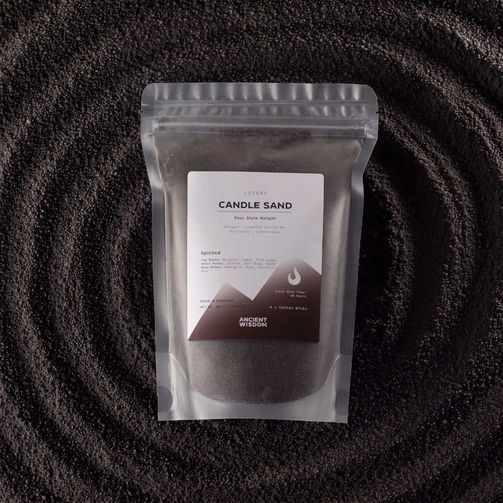 A stand-up pouch of Luxurys Candle Sand (Pura Spyle Recipe) by Ancient Mission, filled with dark gray wax, set against a dark textured background with concentric circular patterns.