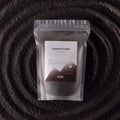 A stand-up pouch of Luxurys Candle Sand (Pura Spyle Recipe) by Ancient Mission, filled with dark gray wax, set against a dark textured background with concentric circular patterns.