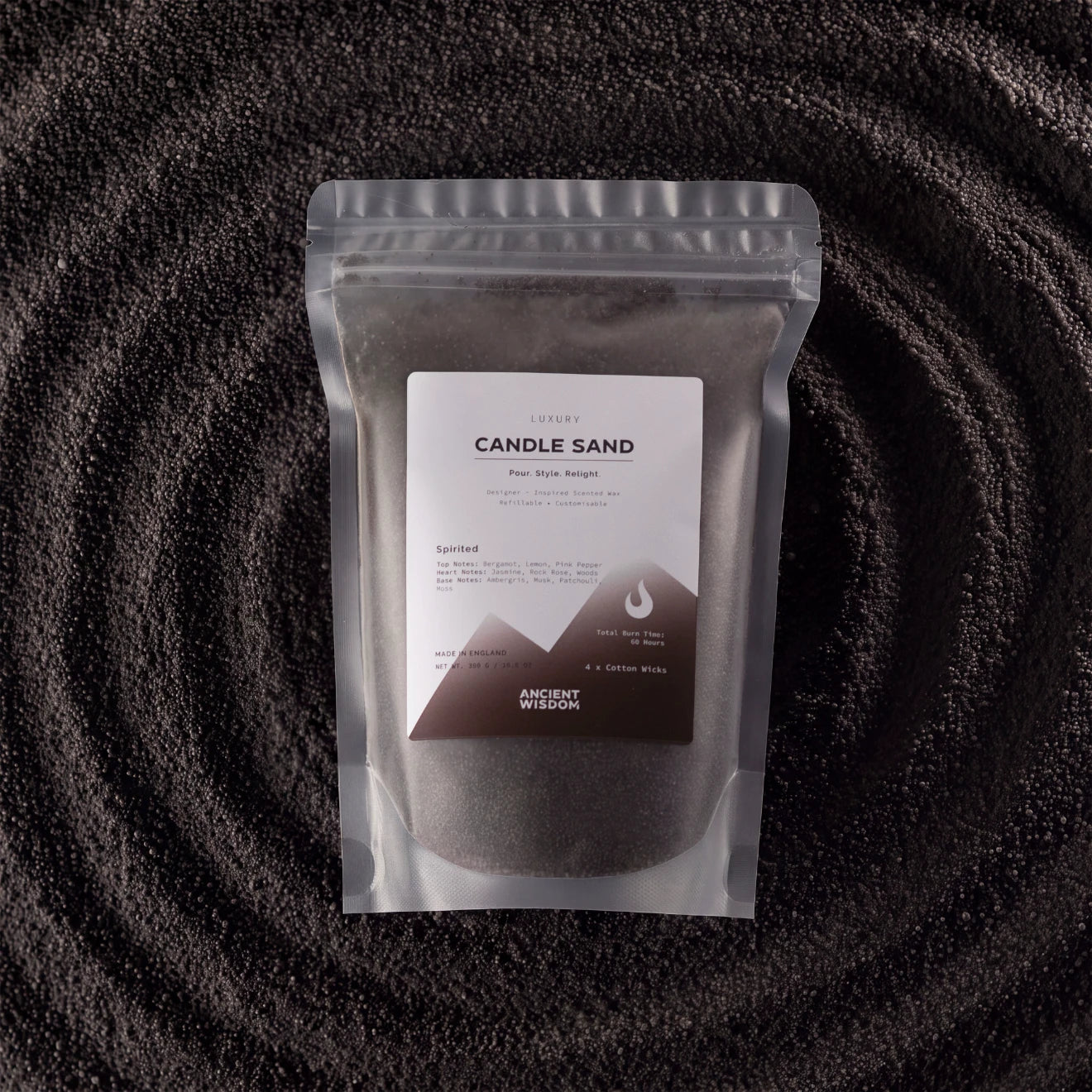 A stand-up pouch of Luxurys Candle Sand (Pura Spyle Recipe) by Ancient Mission, filled with dark gray wax, set against a dark textured background with concentric circular patterns.