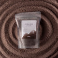 A stand-up pouch labeled 'Candle Sand' filled with dark brown wax, placed on a textured brown surface with concentric circular patterns.