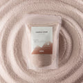 A clear stand-up pouch containing fine beige candle sand, centered on a matching beige powder background with concentric circular texture. The front label reads 'CANDLE SAND' and 'ANCIENT WISDOM'.