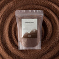 A stand-up pouch of Ancient Wisdom Candle Sand (Flour Size Weight) placed on top of a circular arrangement of the same brown wax-like product, creating a textured background.