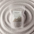 A clear resealable pouch containing white wax melts or wax beads, with a white label reading 'CANDLE SAND' and 'ANCIENT WISON', placed on a textured white fabric background with concentric circular ripples.