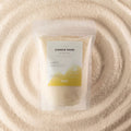 A stand-up pouch labeled 'CANDLE SAND' containing pale cream wax, placed on a textured surface of matching cream wax in a circular pattern. The label has a white top half and a yellow-green geometric bottom section.