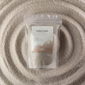 A clear stand-up pouch labeled 'Candle Sand' containing a light beige textured powder, placed on top of concentric rings of the same powder. The pouch has a white and taupe label with the brand name Ancient Wisdom visible near the bottom.