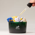 Green container with dried flowers and a dropper adding liquid, featuring the Illo Villare brand.