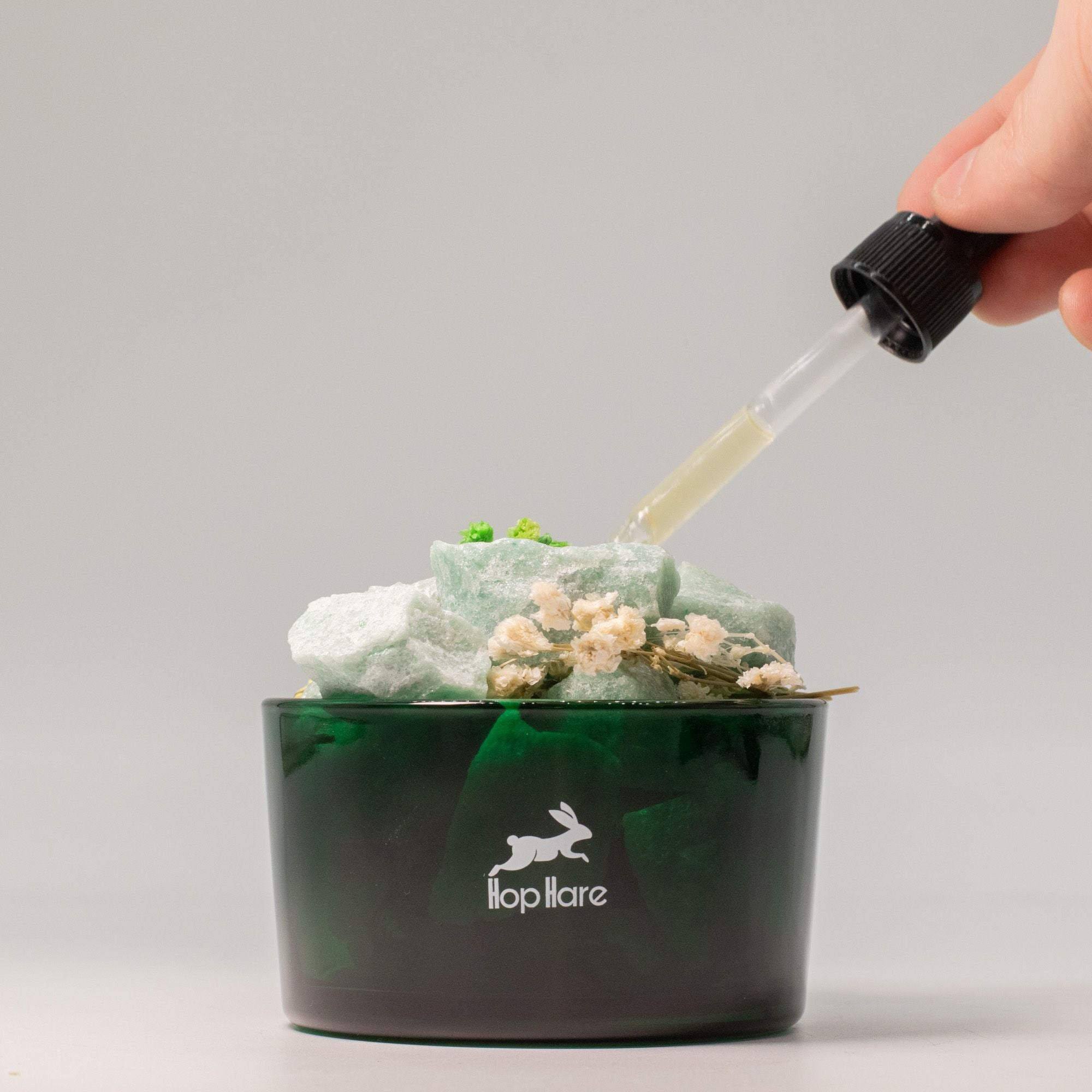 Hand using a dropper Hop Hare Diffusing Crystals & Floral Set to add liquid to a green container with a brand logo on a gray background